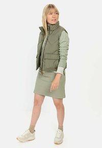 Olijfgroene geïsoleerde vest met hoge kraag, ritsluiting en voorzakken, gelaagd over een lichtgroen topje en gecombineerd met een groene rok en sneakers.