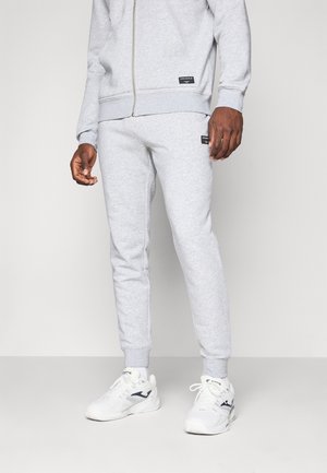 Uomo che indossa pantaloni della tuta grigio chiaro, felpa con cappuccio e zip abbinata, scarpe sportive bianche, in piedi su uno sfondo bianco semplice.