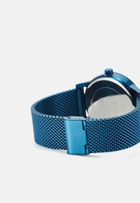 Correa de reloj de malla metálica azul con caja redonda pulida en plata y un cierre grabado con el logo. Textura suave y diseño elegante.