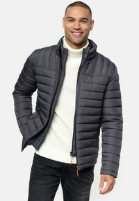 Chaqueta acolchada gris con cremallera frontal, cuello alto y bolsillos laterales; presenta paneles texturizados y tiradores de cremallera de color naranja. Lleva puesta sobre un suéter de punto blanco.