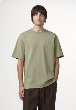 Olijfgroene katoenen t-shirt met korte mouwen, relaxed fit en subtiele voorborduring. Gecombineerd met een bruine broek.