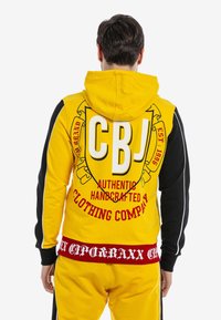 Kapuzensweatshirt in leuchtendem Gelb mit schwarzen Ärmeln. Auf der Rückseite befindet sich große rote Schrift und Logos. Hergestellt aus weichem Stoff.