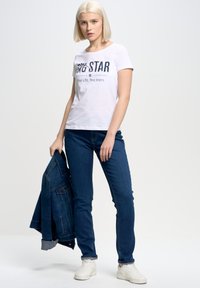 Bílá krátká trička s krátkým rukávem s nápisem "BIG STAR", kombinovaná s tmavě modrými džíny a bílými teniskami. Džínová bunda držena v levé ruce.