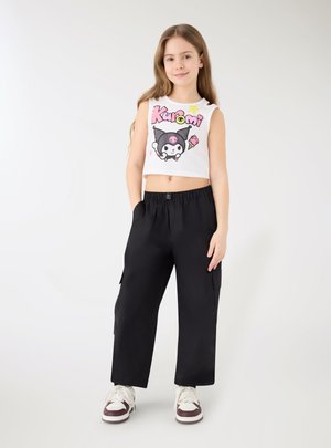 Ragazza giovane con pantaloni neri a gamba larga, crop top bianco e rosa con disegno cartoon, e scarpe da ginnastica bianche, in piedi con una mano in tasca su sfondo bianco.