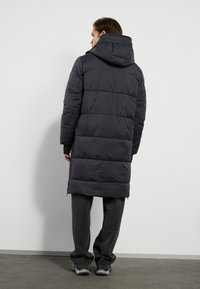 Doudoune grise avec capuche, tissu texturé, longueur longue, coutures horizontales, zips sur les côtés, portée sur un pantalon gris et des baskets.