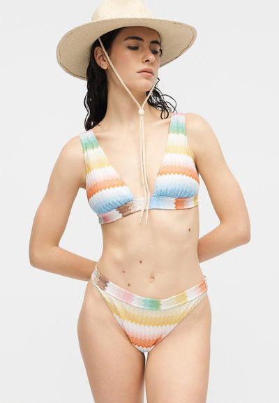 Femme portant un chapeau beige à large bord et un bikini rayé multicolore avec des motifs en zigzag, debout devant un fond blanc uni.
