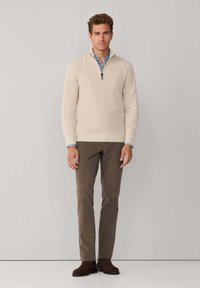 Beige gebreide trui met een kwart-rits kraag, gecombineerd met een paar bruine broek en donkerbruine suède laarzen, model staat tegen een neutrale achtergrond.