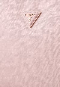 Materiale sintetico rosa testurizzato con un logo triangolare dorato che presenta "GUESS" e "USA" in lettere in rilievo, accentuato da un punto interrogativo.