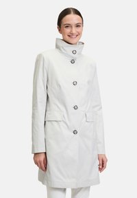 Lichtgrijs, knielang jasje met een hoge kraag, knoopsluiting aan de voorkant en twee zijzakken. Kenmerkt zich door een gladde stof en vijf decoratieve knopen.