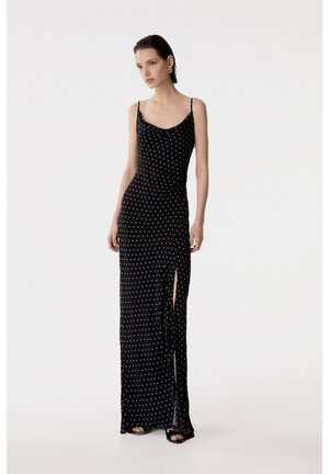 Donna che indossa un abito lungo nero con pois bianchi e uno spacco alto sulla coscia, in piedi davanti a uno sfondo bianco uniforme.