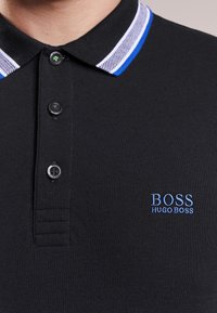 Black polo shirt featuring a textured collar with blue and white stripes. Two button placket with skull detail. Logo embroidered in light blue.