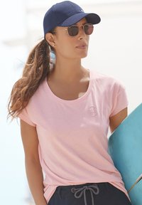 Lichtroze T-shirt met korte mouwen en een ronde halslijn, met een klein geborduurd logo. De drager heeft een marineblauwe pet en een zonnebril.