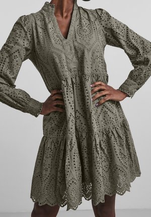 Femme portant une robe en dentelle ajourée vert olive à manches longues, ourlet festonné, col en V et taille froncée, mains sur les hanches.