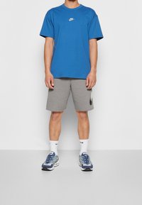 Person som bär en blå Nike t-shirt, grå Nike shorts, vita Nike strumpor och blå och vita Nike Air Max sneakers, stående mot en enfärgad bakgrund.