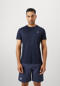 Άνδρας που στέκεται μπροστά σε απλό φόντο, φορώντας ναυτικό αθλητικό μπλουζάκι και σορτς με μικρά λευκά λογότυπα στο μπλουζάκι και στο αριστερό πόδι του σορ�τς.