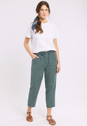 Jeune femme aux longs cheveux attachés avec un foulard bleu, portant un t-shirt blanc, un pantalon vert à cordon et des sandales marron, debout, une main dans la poche.