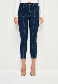 Džínsy s vysokým pasem, tmav ého denimu, s kontrastním šitím, slim fit, zkrácené délky a předními kapsami. Nosí se s nahými vysokými podpatky.