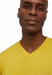 Gelbes V-Ausschnitt-Pullover aus weichem Strickstoff mit strukturierter Oberfläche und sauberem Ausschnitt, teilweise von einem Model getragen.