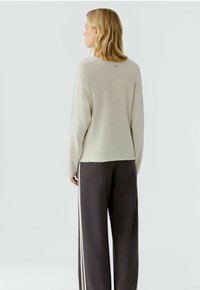 Beiger Pullover mit langen Ärmeln, Rundhalsausschnitt und einem kleinen Akzentdetail am Rücken; kombiniert mit dunkelgrauen Weitbein-Hosen mit weißen Seitenstreifen.