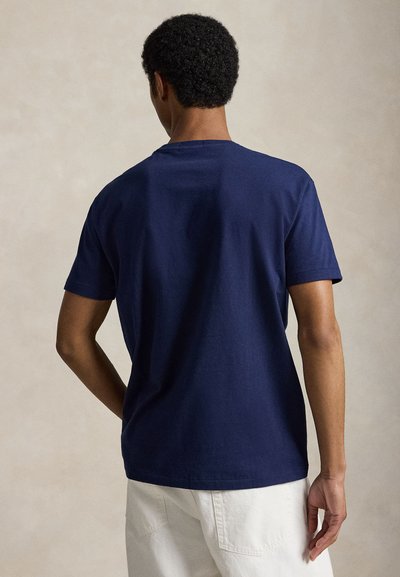 Άνδρας με κοντά, σγουρά μαλλιά που φοράει ένα μονόχρωμο σκούρο μπλε κοντομάνικο μπλουζάκι και λευκό παντελόνι, στέκεται πλάτη προς τα εμπρός.