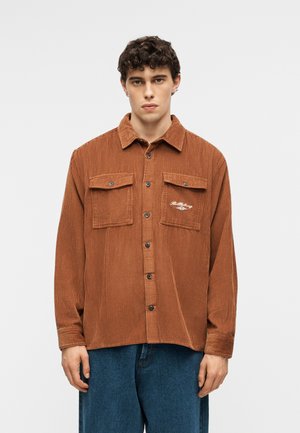 Νεαρός άνδρας που φοράει ένα καφέ πουκάμισο κοτλέ με κουμπιά και πλαϊνές τσέπες με καπάκι, και μπλε τζιν, στέκεται μπροστά σε ένα απλό φωτεινό φόντο.