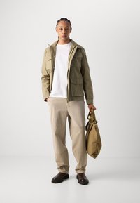 Jachetă de un verde măsliniu deschis cu multiple buzunare frontale, tricou alb, pantaloni bej și cizme maro închis. Poartă o geantă bej cu bretele pentru umăr.
