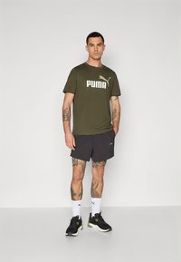 Õlivärvi rohelise Puma t-särk valge logoga, mida kombineeritud mustade lühikestega. Modell kannab musti tossusid ja valgeid sokke Puma logoga.