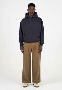 Maritieme capuchontrui met voorzak, gecombineerd met bruine oversized broek en crème schoenen. Model staat tegen een plain witte achtergrond.