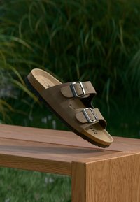 Sandalo in suede marrone con due fibbie regolabili in metallo argento, plantare sagomato e suola nera testurizzata, posizionato su una superficie in legno.