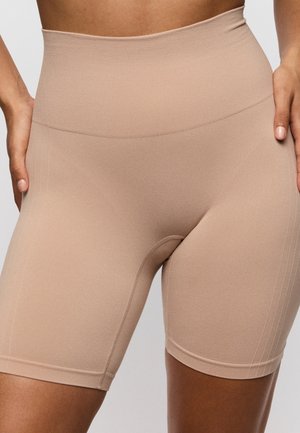 Close-up van een persoon die beige, naadloze biker shorts met hoge taille draagt, met handen op de heupen tegen een effen achtergrond.