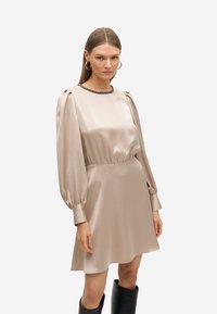 Vestido de satén color beige claro con mangas abullonadas, cintura ajustada y escote redondo adornado con ribete negro. Textura suave, con dobladillo a la altura de la rodilla.