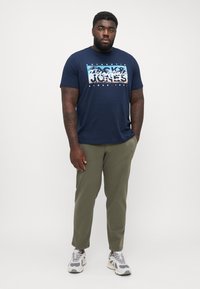 Mørkeblå t-shirt med korte ærmer og grafisk print med hvid tekst, parret med olivengrønne bukser og grå sneakers med hvide accenter.
