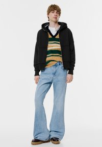 Fekete zipzáras kapucnis pulóver, egy puha csíkos pulóver felett, ami barna, zöld és fehér színekben pompázik. Világoskék bővülő farmernadrág és vastag barna cipők.