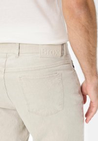 Light beige denim trousers with a smooth texture, featuring a Brax logo patch on the waistband and one rear pocket with a subtle design.