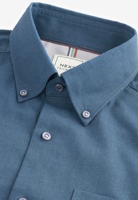 Camisa azul texturizada de botón con cuello clásico, que presenta botones claros y una etiqueta con rayas multicolores en el interior del cuello.