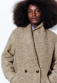 Manteau beige à chevrons croisé avec un large col, tissu texturé et grands boutons foncés ; conçu avec des poches latérales.