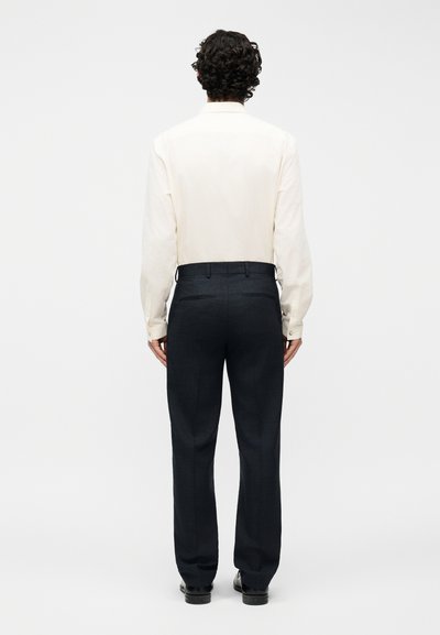 Άνδρας με σγουρά σκούρα μαλλιά, γυρισμένος πλάτη, φοράει μακρυμάνικο πουκάμισο εκρού, μπουσουρισμένο σε μαύρο παντελόνι προσαρμοσμένης γραμμής και μαύρα παπούτσια.