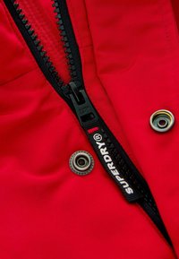 Röd tyg med en svart dragkedjestängning, med en texturerad yta, metallknäppning och en framträdande "SUPERDRY"-etikett.
