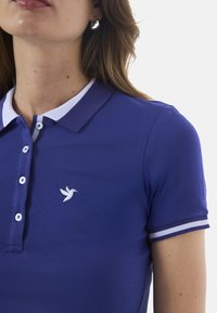 Fioletowa koszulka polo z białym kołnierzykiem, zapięciem na trzy guziki, haftowanym logo ptaka i paskowymi akcentami na rękawach. Gładka tekstura.
