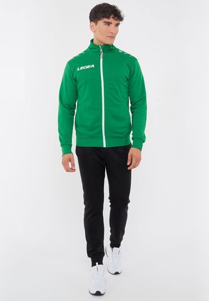 Man wearing green zip-up sports jacket, black pants, and white sneakers, standing and looking to the side on a white background.