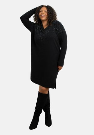 Zwarte oversized sweaterdress met een V-halslijn en versierde pailletten op de schouders. Zachte textuur, knielang met zijsplitten.