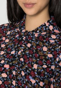 Svart blommig blus med små rosa, röda och blå blommor, klassisk krage, knappfram och mjuk tygtextur.