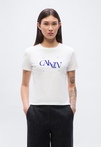 Λευκό βαμβακερό μπλουζάκι με έντονο μπλε κείμενο "CALVIN KLEIN NEW YORK." Το μοντέλο φοράει μαύρο, υφασμάτινο παντελόνι φαρδιάς γραμμής.