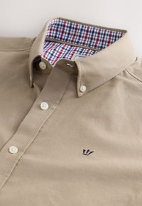 Beige Hemd mit Knopfleiste, geknöpfter Kragen, weißen Knöpfen, marineblauem Kronenlogo auf der Brust und einem rot-weiß-marineblauen karierten Innenkragen.