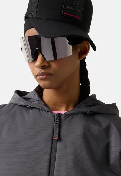 Osoba w czarnej czapce, dużych odblaskowych sportowych okularach przeciwsłonecznych oraz szarej kurtce z kapturem na zamek, zwrócona nieco w lewo na białym tle.