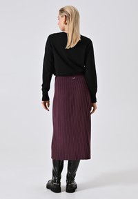 Donna con capelli biondi che indossa un top nero a maniche lunghe, una gonna midi viola a coste e stivali neri al ginocchio, vista di spalle su uno sfondo neutro.