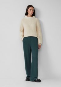 Pull crème épais avec un col roulé haut, associé à un pantalon large vert foncé. Le mannequin se tient contre un fond neutre.