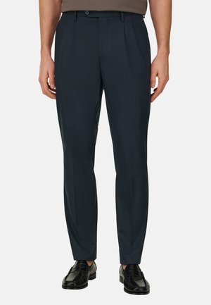 Uomo che indossa pantaloni eleganti blu scuro e mocassini di pelle nera, in piedi con le mani rilassate lungo i fianchi su uno sfondo bianco.