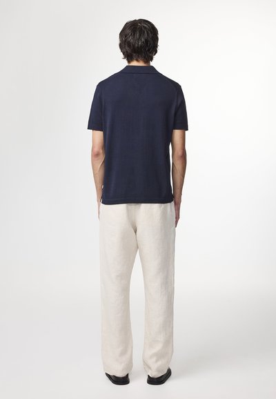 Mann steht mit dem Rücken zur Kamera, trägt ein marineblaues Poloshirt mit kurzen Ärmeln, hellbeige Hosen und schwarze Schuhe vor einem weißen Hintergrund.