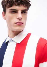 Νεαρός με σγουρά μαλλιά που φοράει μπλουζάκι πόλο με ρίγες κόκκινες,  λευκές και μπλε, κοιτάζει ελαφρώς προς τα πλάγια με φόντο ένα λευκό τοίχο.